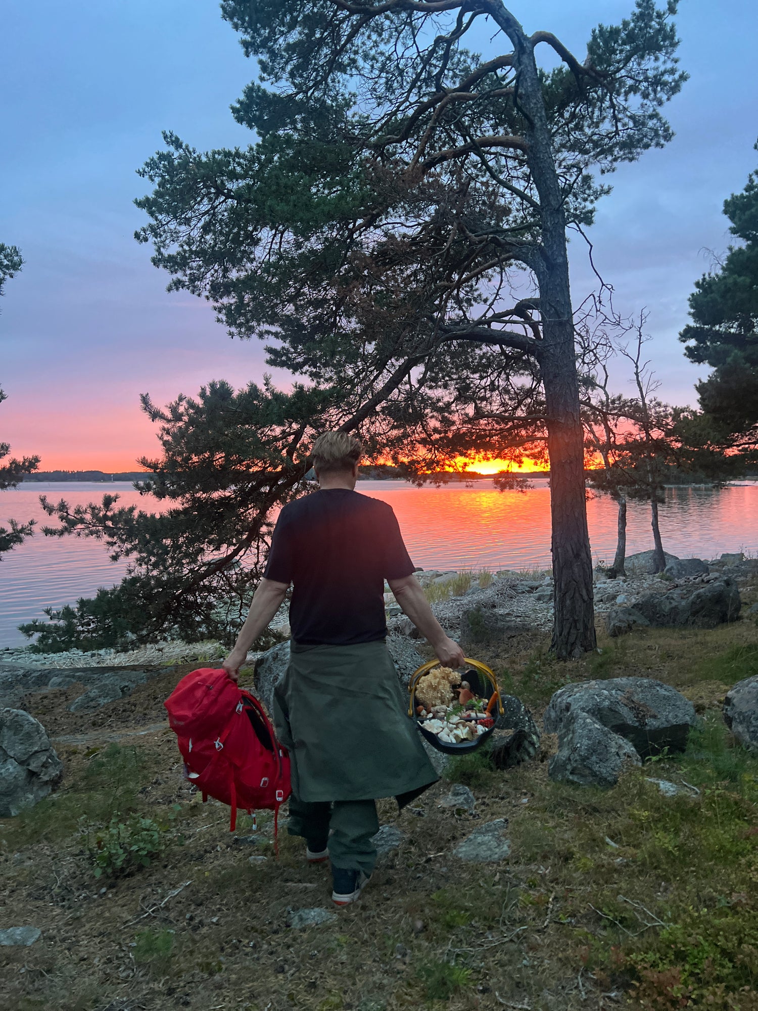 Henkilö kantaa punaista reppua ja koria järven rannalla auringonlaskun aikaan, etualalla puita ja kiviä.