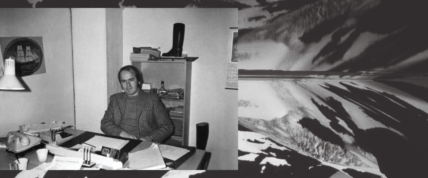 Mies istuu toimistossa sekavan työpöydän ääressä; hänen takanaan hyllyllä on saappaat.