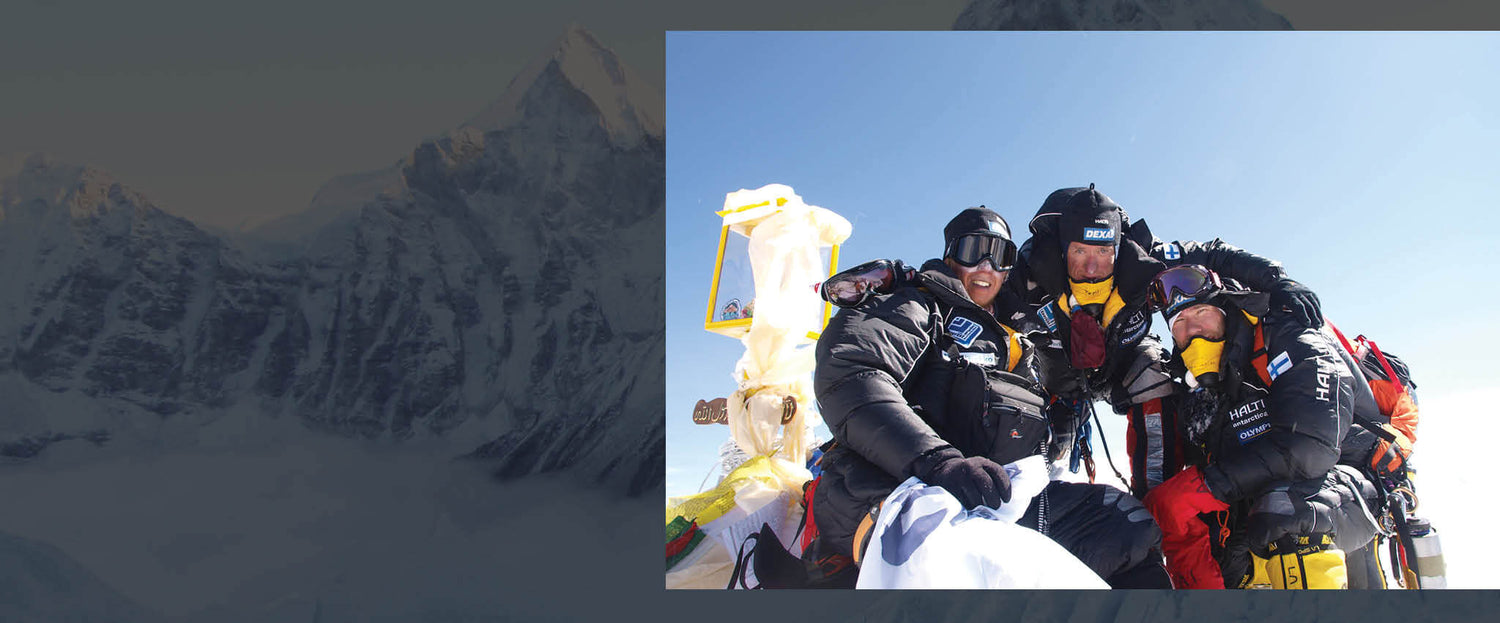 Kolme kiipeilijää raskaissa varusteissa syleilee ja hymyilee lumisella vuoren huipulla kirkkaana, aurinkoisena päivänä.