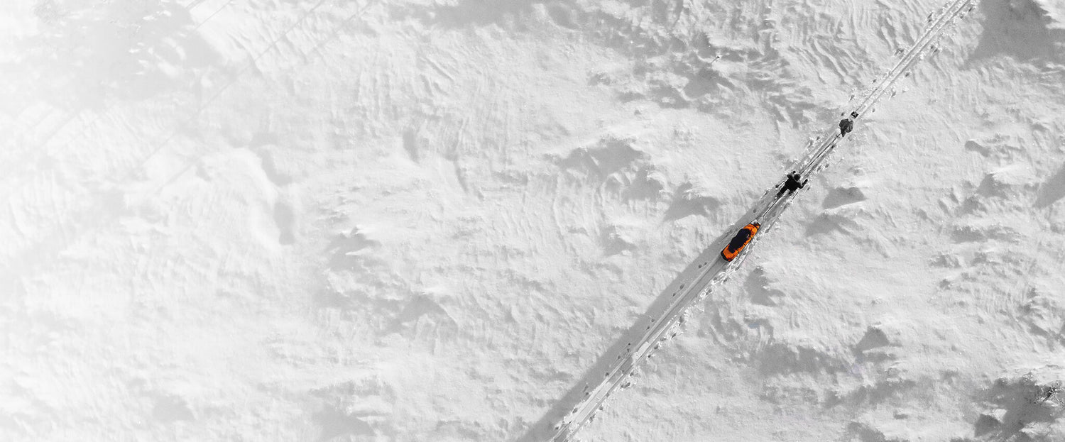 Pieni oranssi auto ajaa lumipeitteisellä tiellä, ja kaksi ihmistä kävelee lähistöllä ylhäältä katsottuna.