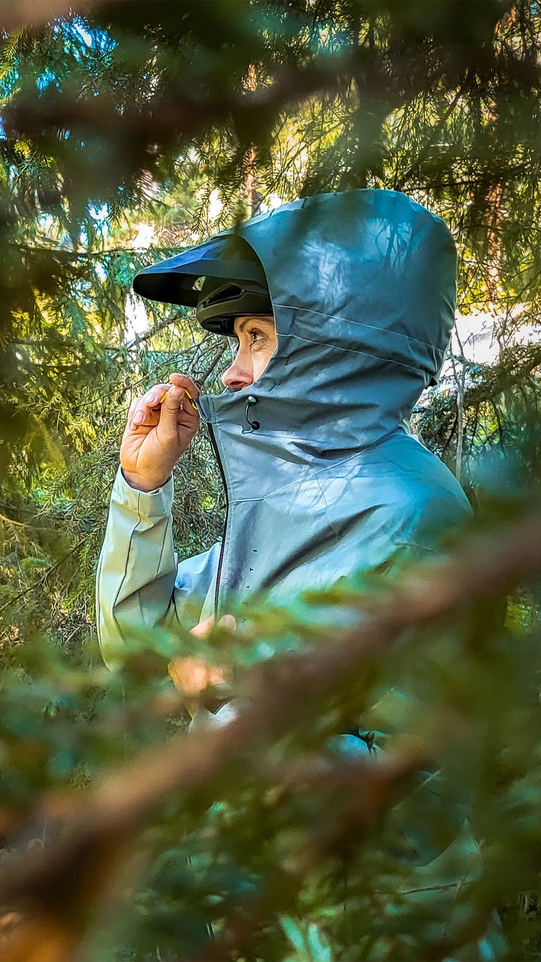 Harmaaseen sadetakkiin ja kypärään pukeutunut henkilö seisoo puiden välissä, vetää huppunsa ylös ja on osittain oksien peitossa.