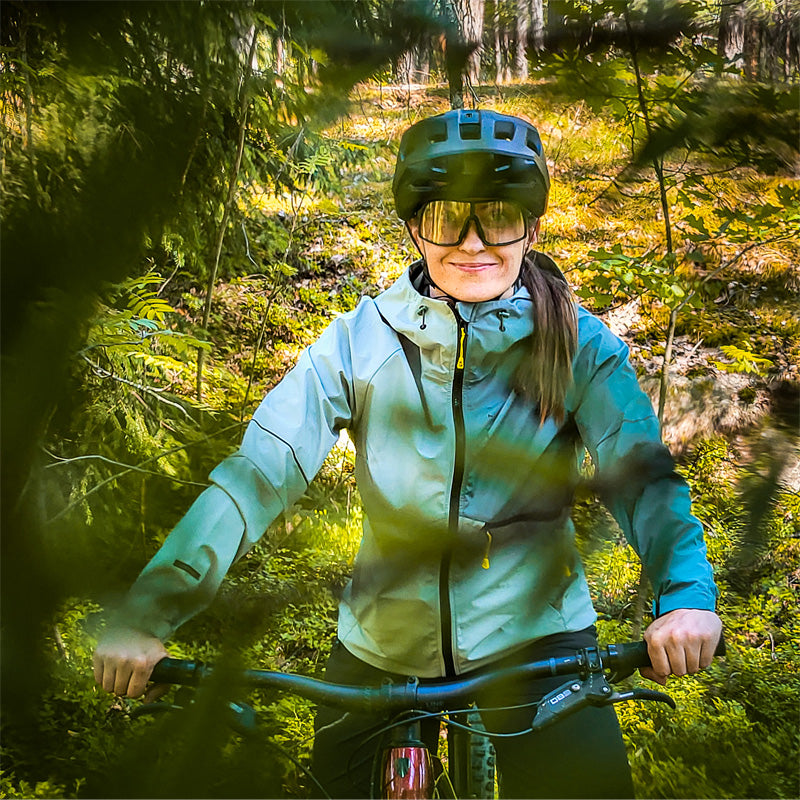 Henkilö, jolla on kypärä ja aurinkolasit, ajaa polkupyörällä metsäisellä alueella, jossa on vihreää lehtimetsää.