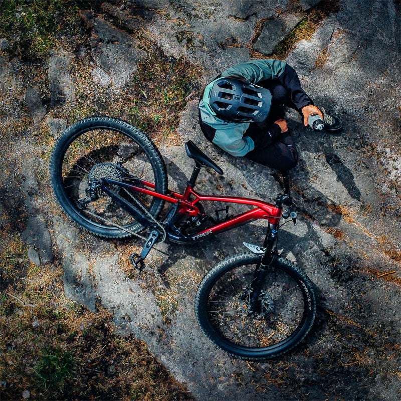 Kypäräpäinen henkilö istuu kivisellä maalla punaisen maastopyörän vieressä ylhäältä katsottuna.