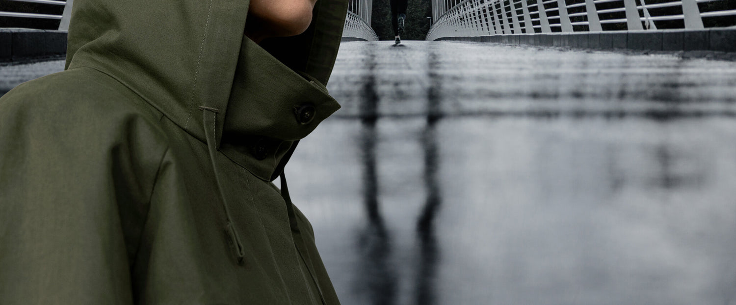 Oliivinvihreään hupulliseen takkiin pukeutunut henkilö seisoo märän, tyhjän sillan vieressä, jossa on valkoiset kaiteet, kasvot kamerasta poispäin.