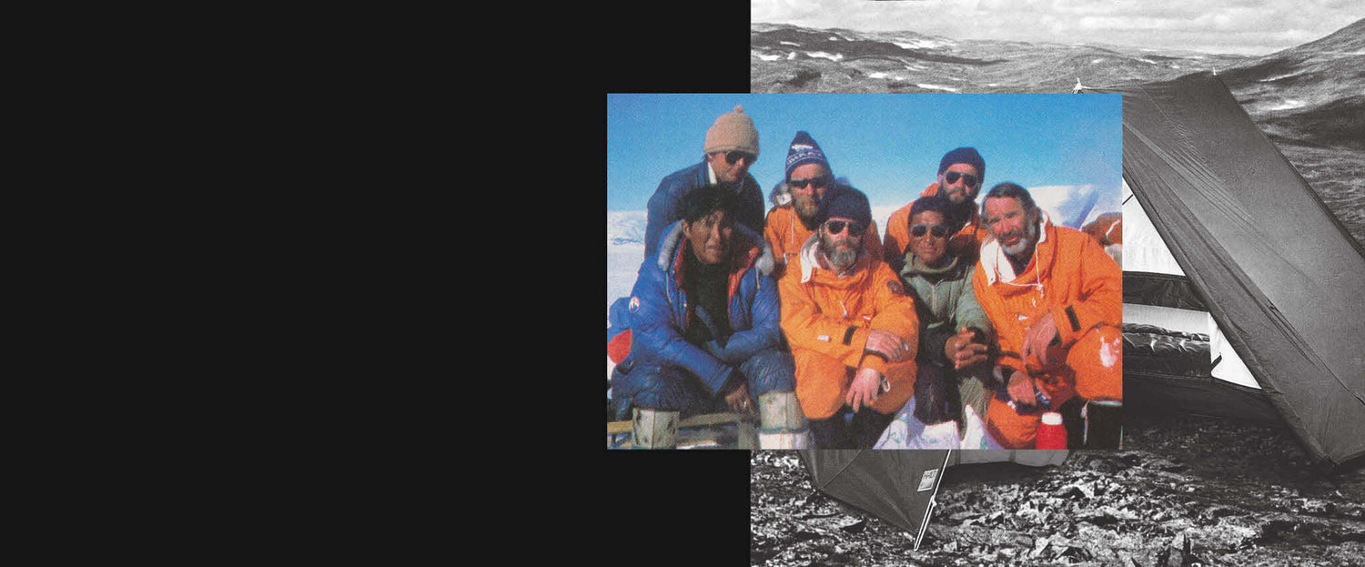 Seitsemän kylmävarusteisiin pukeutuneen henkilön ryhmä poseeraa yhdessä ulkona lumisella vuoristoalueella telttojen läheisyydessä.