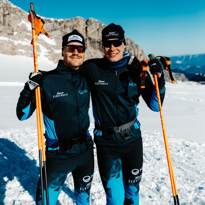 Kaksi talviurheiluasuun pukeutunutta ihmistä seisoo lumella, kädessään oranssit hiihtosauvat, taustalla vuoret.