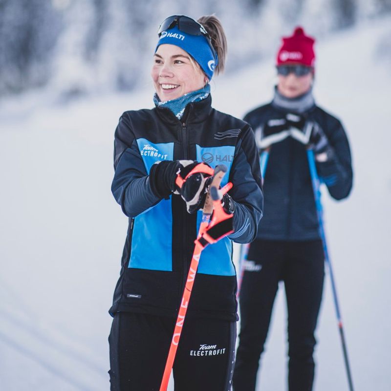 Kaksi ihmistä hiihtämässä talvella, toinen hymyilee etualalla ja taustalla on lumisia puita.