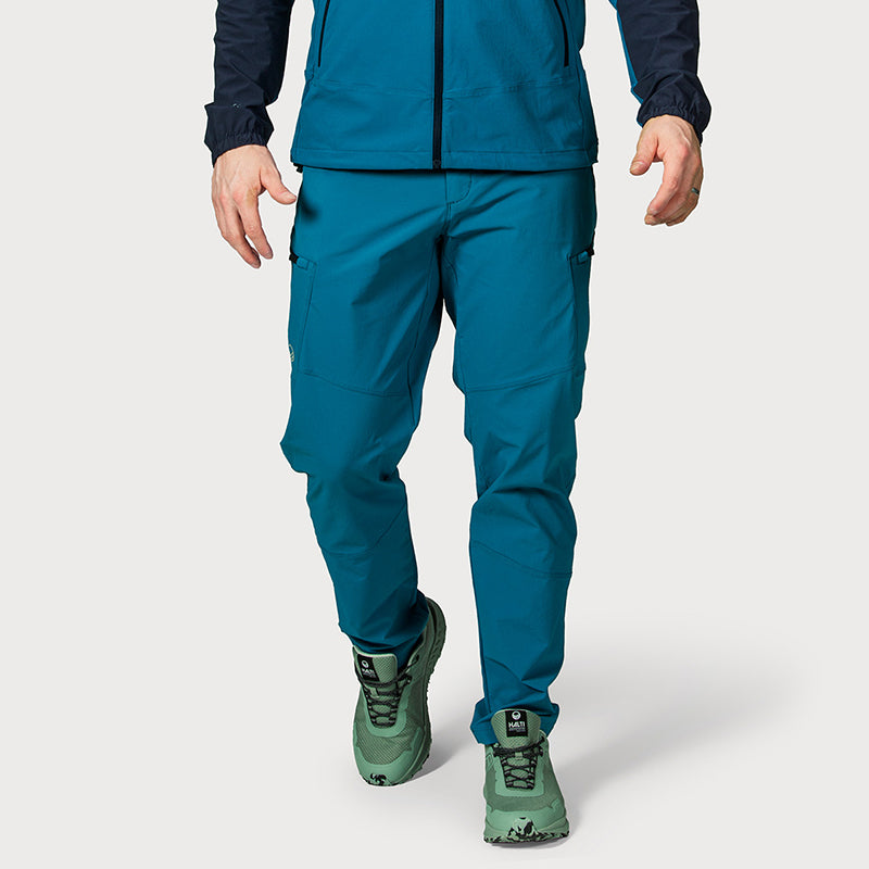 Kuvassa henkilö sinivihreässä Halti Pallas Evo X-stretch -housuissa, yhteensopivassa takissa ja vihreissä maastokengissä olkapäistä alaspäin.