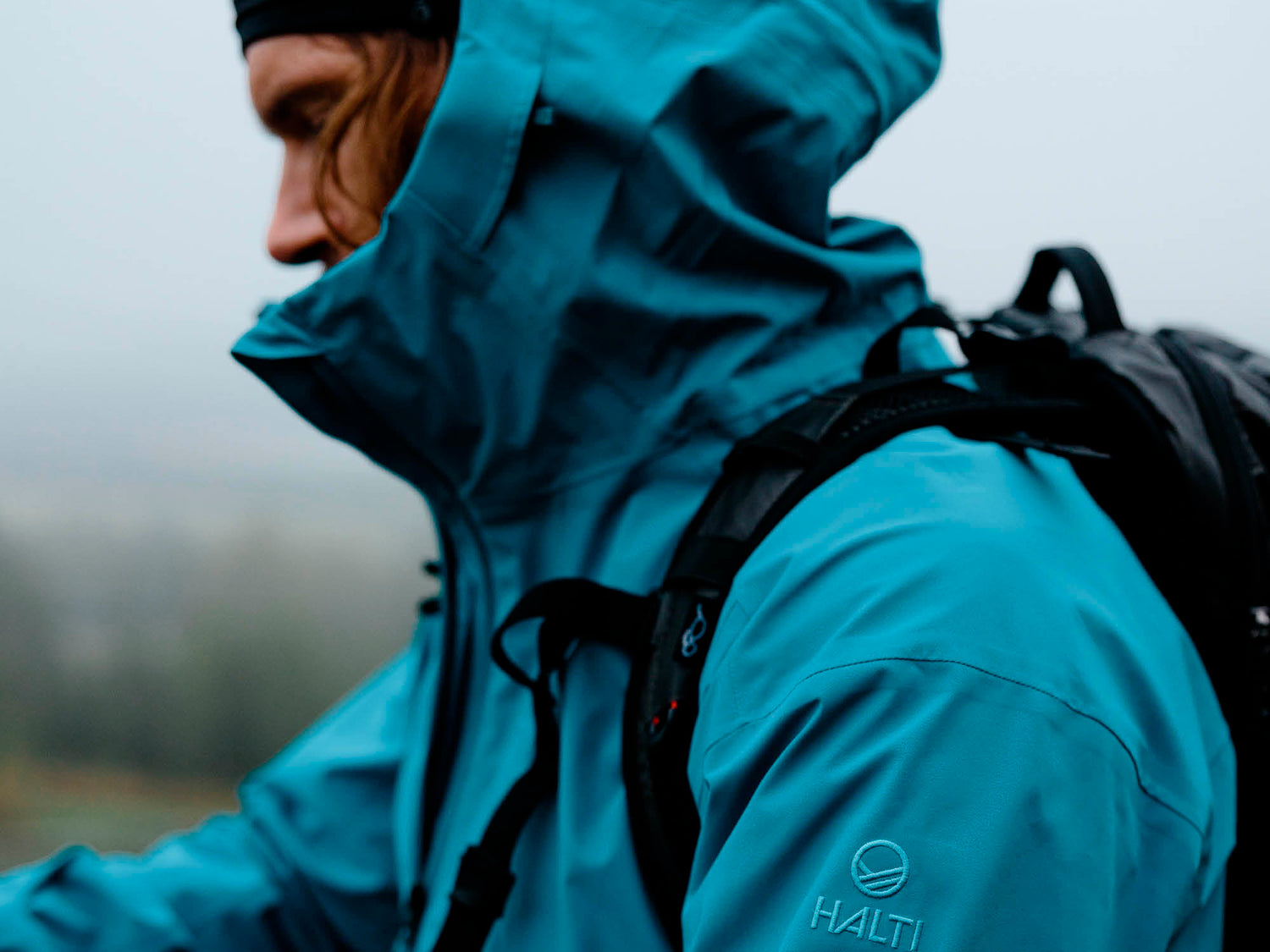 Henkilö, jolla on sininen Halti-takki ja huppu ylhäällä, yllään musta reppu, ulkona sumuisessa ympäristössä.