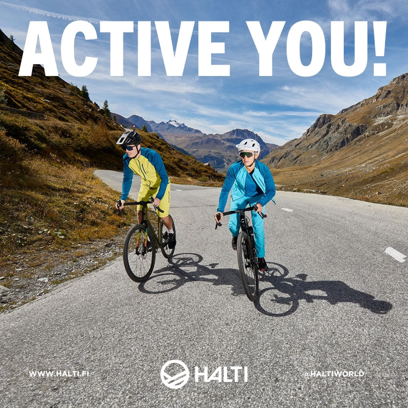 Kaksi pyöräilijää ajaa päällystettyä vuoristotietä sinisen taivaan alla, ja yläreunassa lukee suurilla valkoisilla kirjaimilla