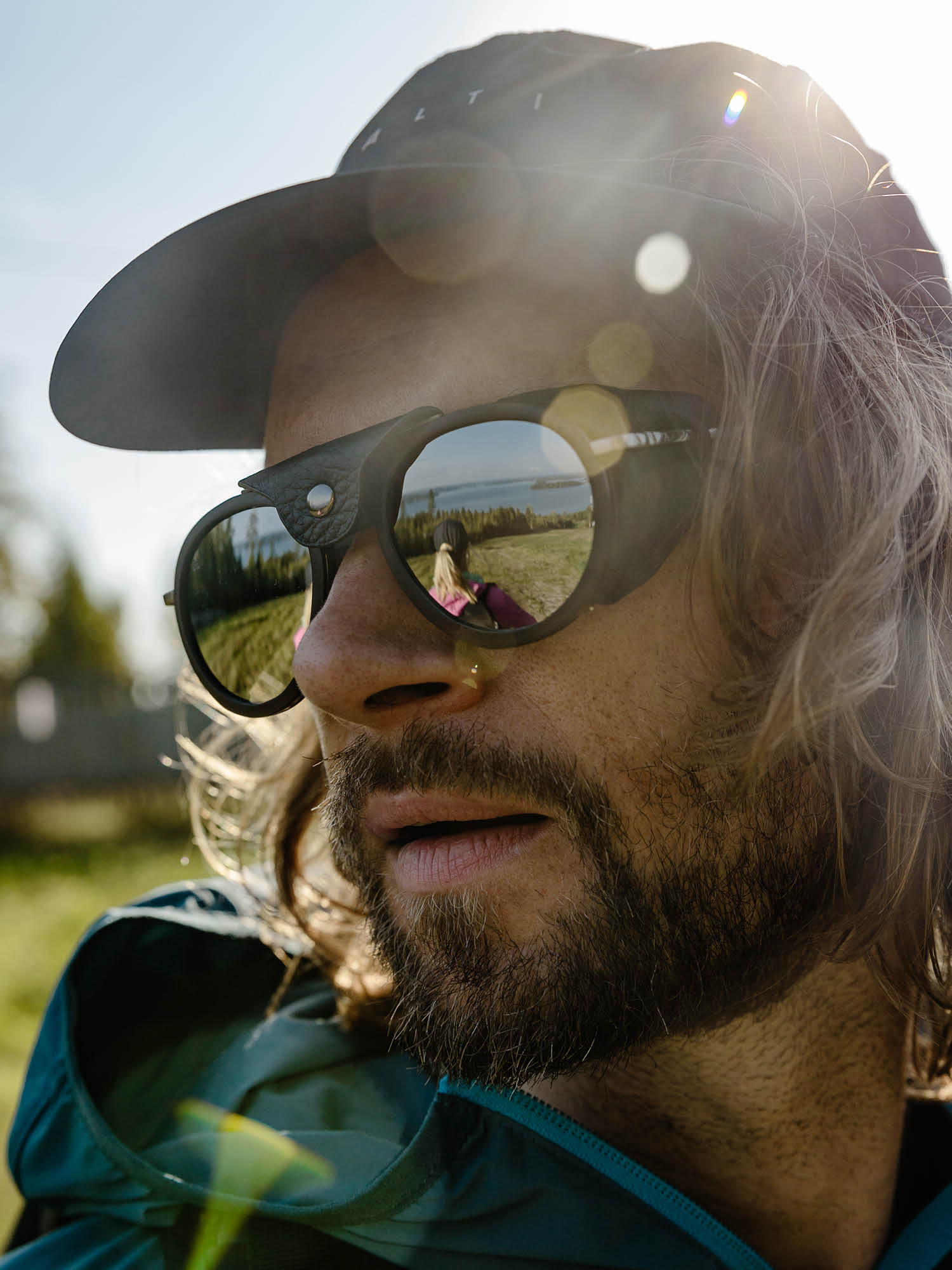 Mies, jolla on aurinkolasit ja lippis ulkona; järvi ja toinen henkilö heijastuvat hänen aurinkolaseissaan.