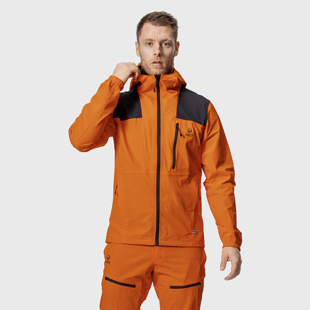 Mies, jolla on oranssi hupparitakki ja housut, joissa on mustat korostukset, seisoo vaaleanharmaata taustaa vasten.