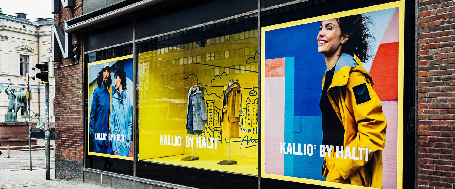 Myymälä, jossa on suuret keltaiset näyteikkunat, joissa on ulkoiluvaatteita ja takkeja käyttäviä malleja; taustalla kaupunkikatu.