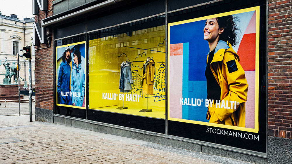 Myymälän näyteikkunassa on Kallio by Haltin vaatteita ja suuria julisteita keltaisia takkeja käyttävistä ihmisistä.