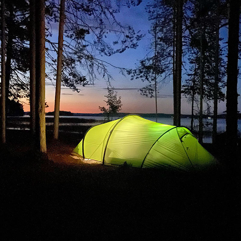 Vihreä teltta on pystytetty puiden keskelle järven lähelle iltahämärässä, ja taustalla on värikäs auringonlaskun taivas.