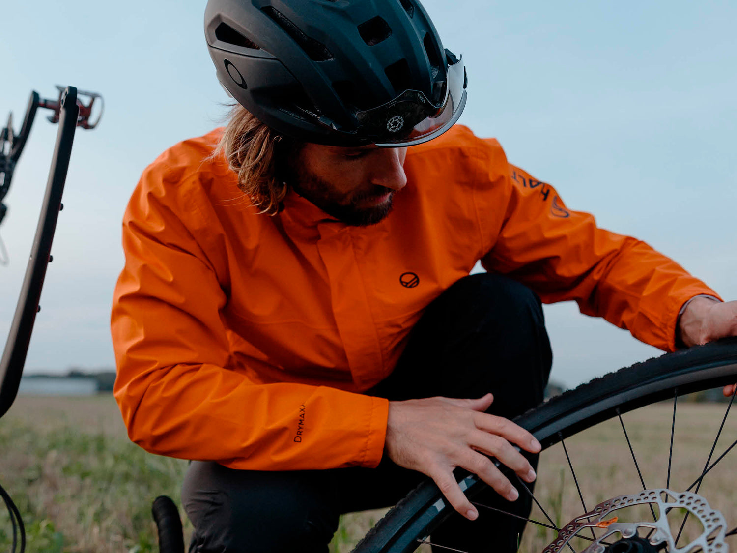 Oranssiin takkiin ja kypärään pukeutunut henkilö tutkii polkupyörän etupyörää ulkona.