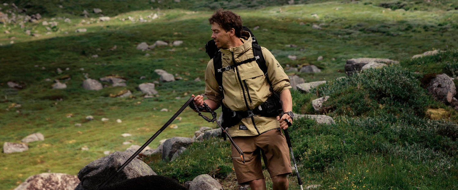 Vaellusvarusteisiin pukeutunut henkilö pitää hihnaa ja vaellussauvoja kädessään seisoessaan ruohikkoisessa, kivikkoisessa maisemassa.