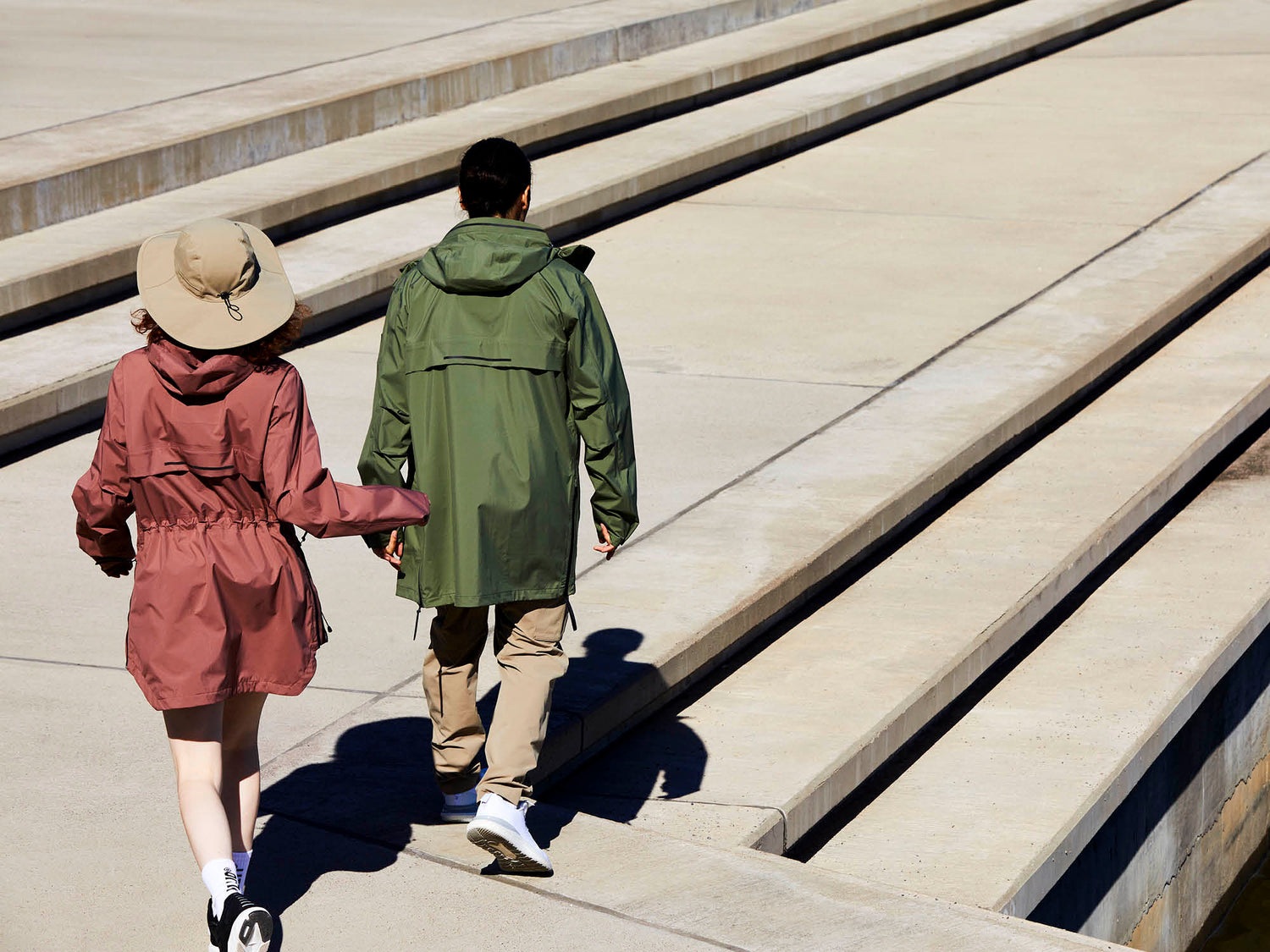 Kaksi ulkoilutakkiin pukeutunutta henkilöä kävelee leveät betoniportaat ylös; toisella on hattu ja punainen takki, toisella vihreä takki.