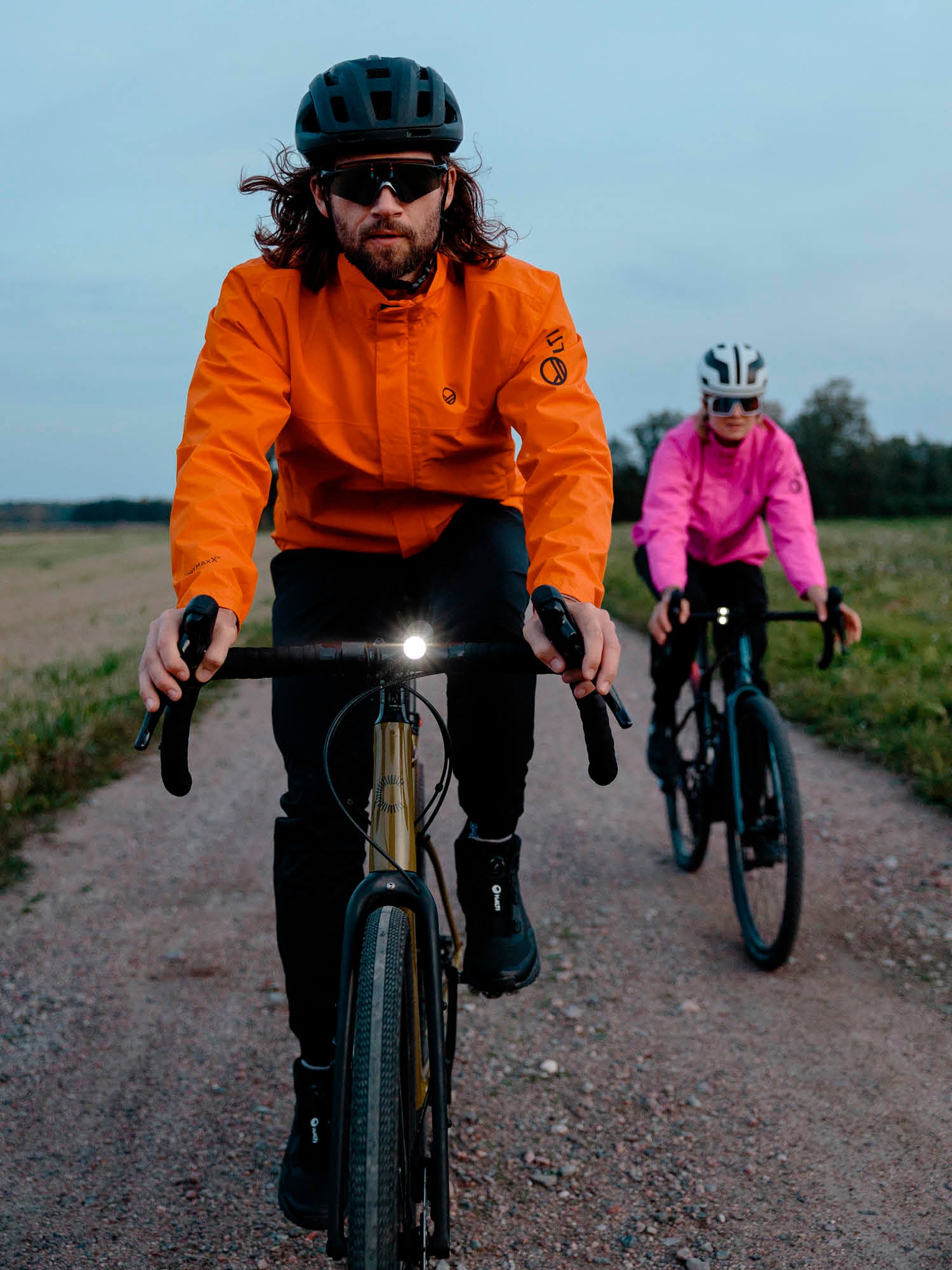 Kaksi pyöräilijää ajaa sorapolkua; toisella on oranssi takki, toisella vaaleanpunainen takki, molemmilla on kypärä ja aurinkolasit.