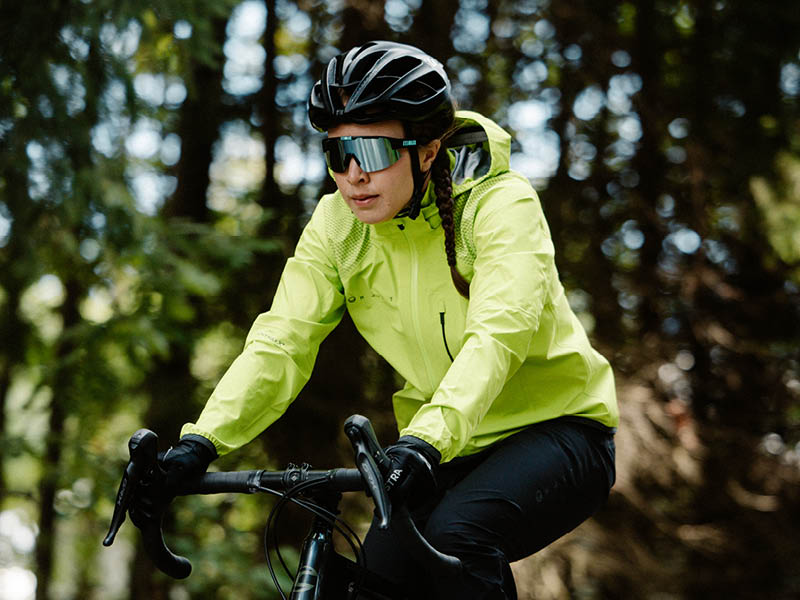 Henkilö, jolla on neonvärinen takki, kypärä ja aurinkolasit, ajaa polkupyörällä metsäisen alueen läpi.