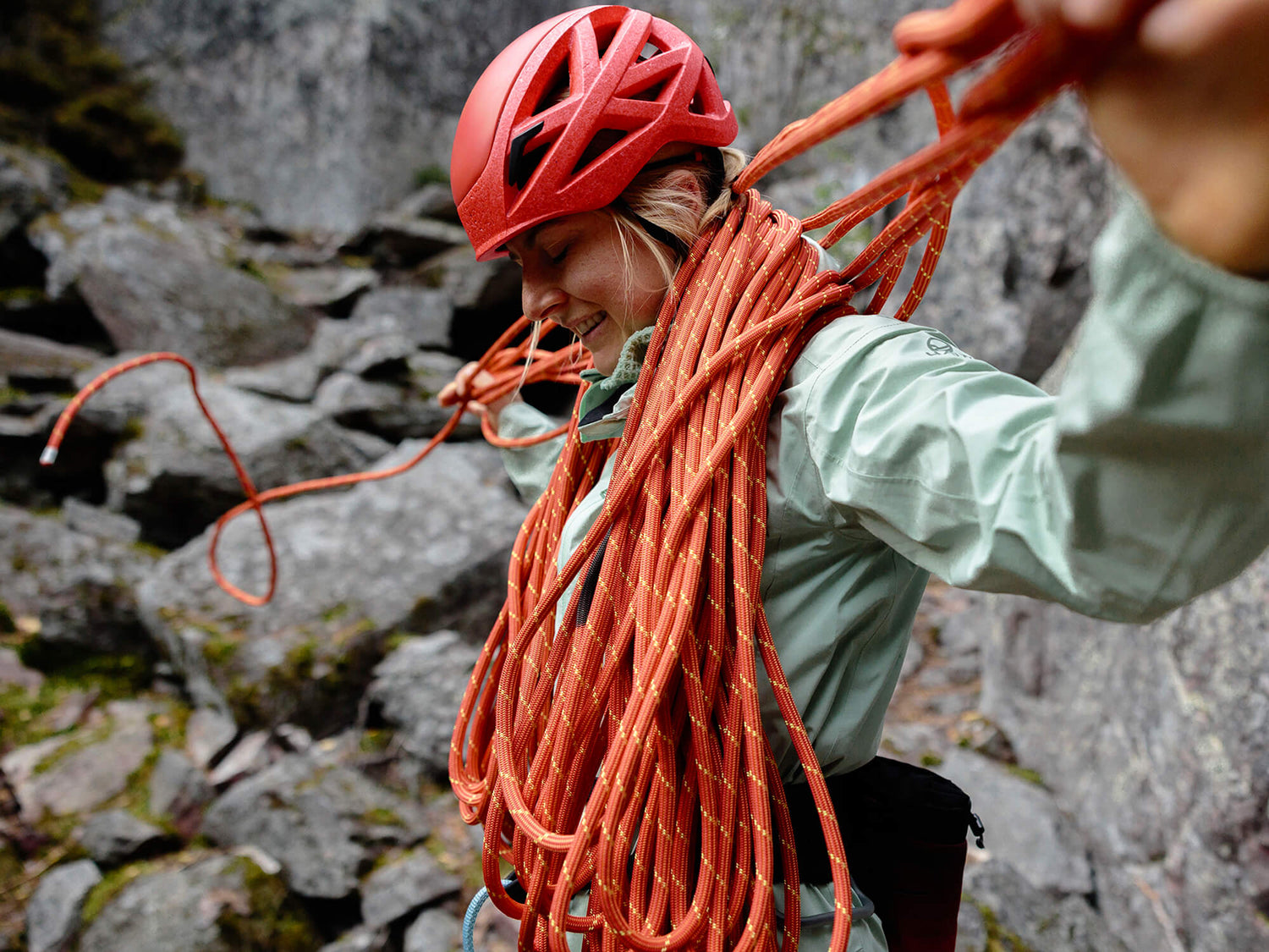 Henkilö, jolla on punainen kypärä ja vaaleanvihreä takki, seisoo ulkona ja kantaa olkapäällään kelattua kiipeilyköyttä.