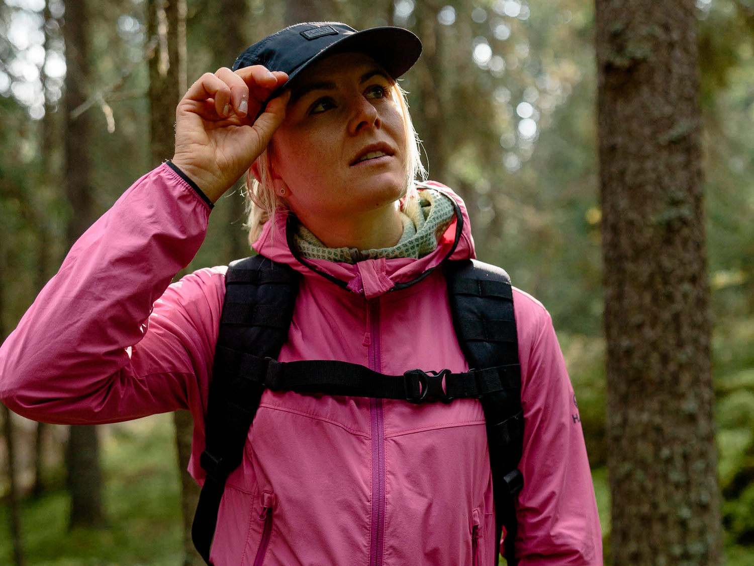 Vaaleanpunaiseen takkiin ja mustaan reppuun pukeutunut henkilö säätää lippalakkia seisoessaan auringon valaisemassa metsässä.
