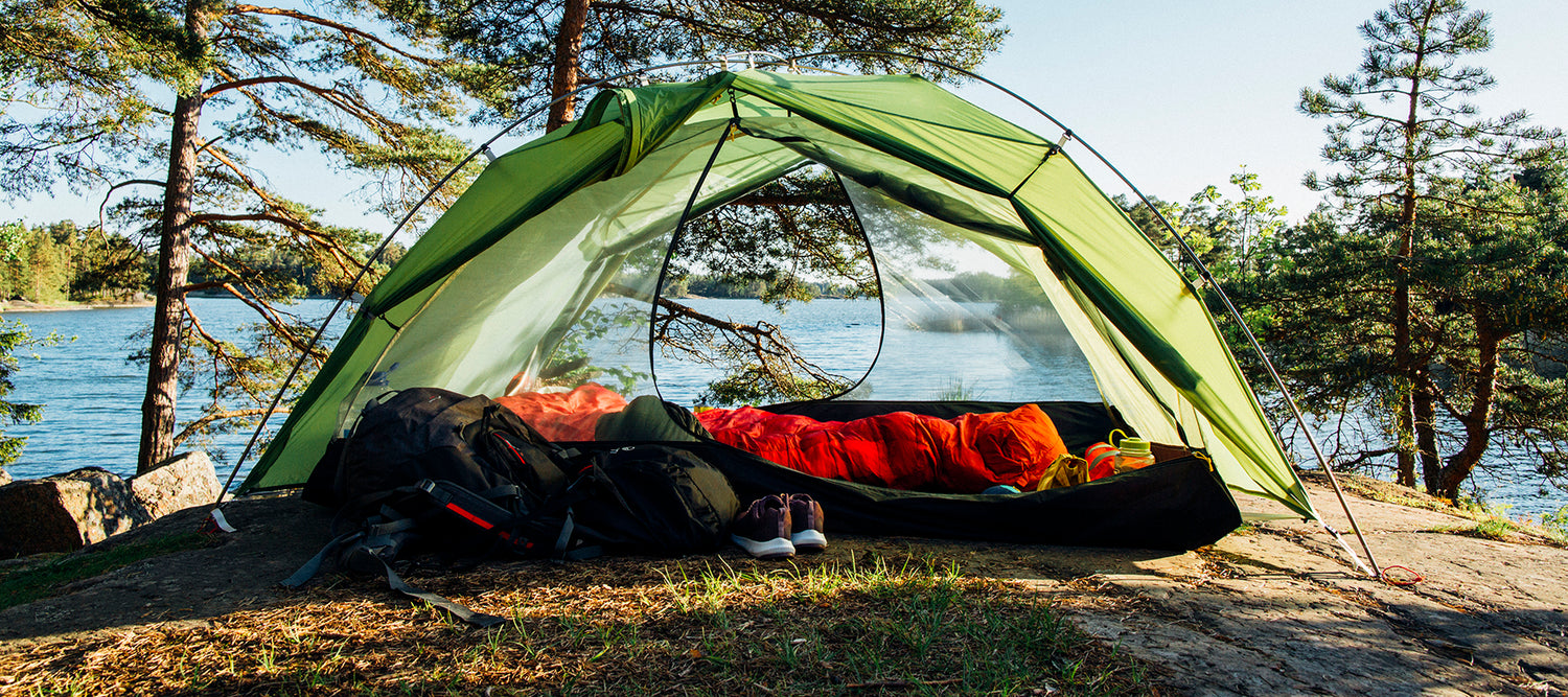 Järven rannalle pystytetty vihreä teltta, jonka sisällä ja ulkopuolella on makuupusseja, reppuja ja kenkiä.