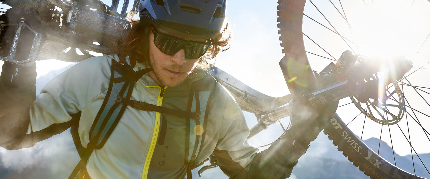 Kypärä ja aurinkolasit päässä oleva henkilö kantaa maastopyörää olallaan ulkona kirkkaassa auringonvalossa.