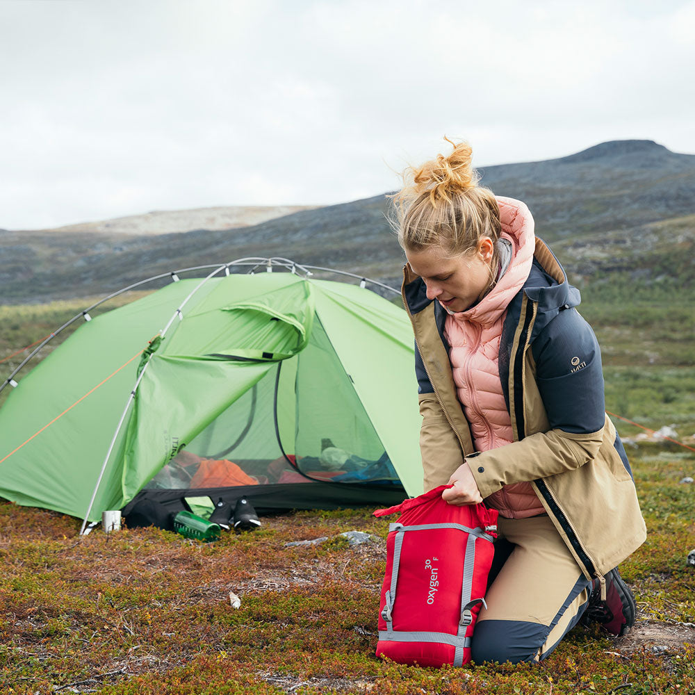 Henkilö polvistuu ruohikolla vihreän teltan vieressä ja pakkaa tavaroita punaiseen pussiin ulkoilmassa vuoristoisella alueella.