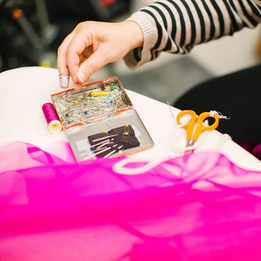 Henkilö valitsee neulan pöydällä olevasta laatikosta, jossa on ompelutarvikkeita ja kirkkaan vaaleanpunaista kangasta.