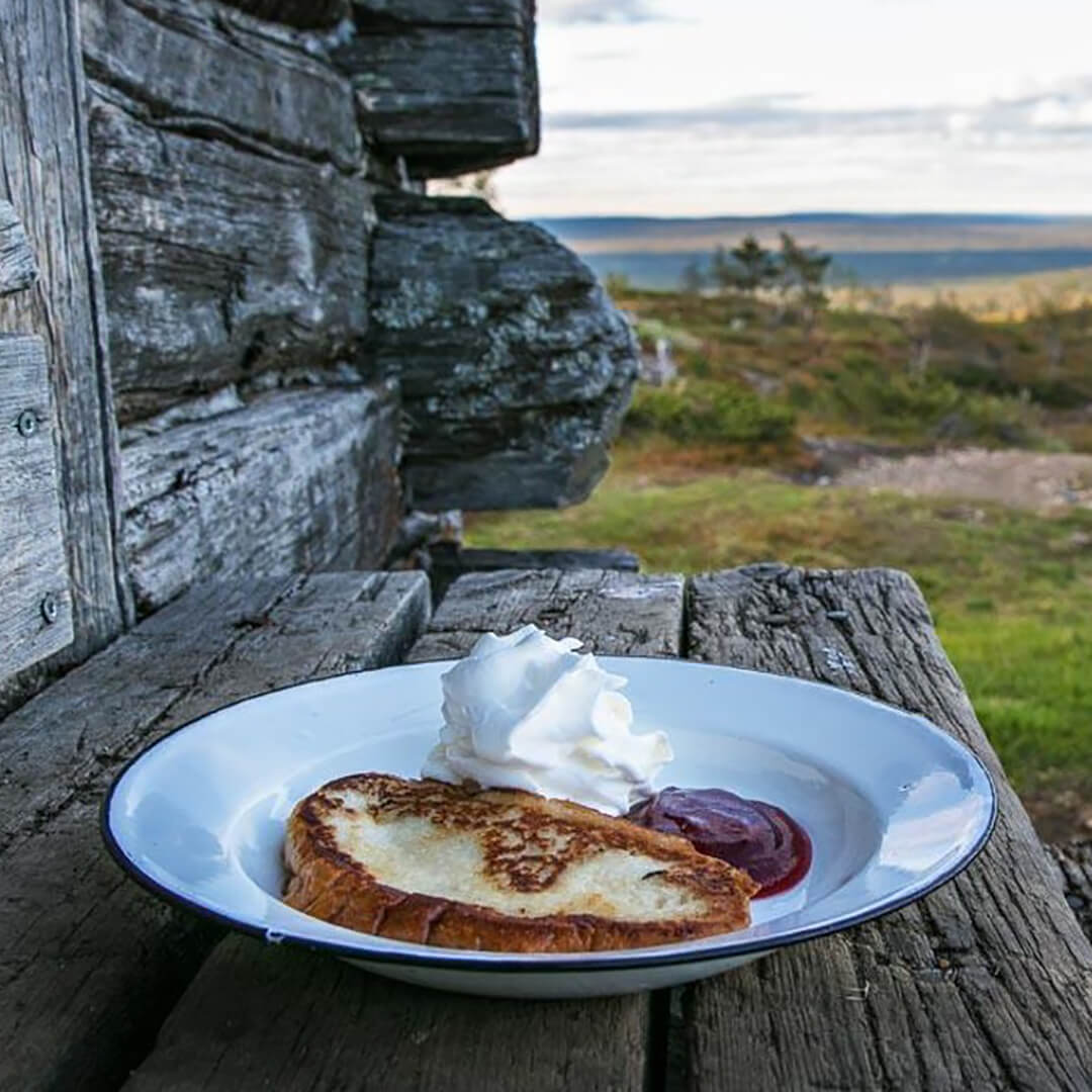 Lautanen, jossa on viipale paistettua leipää, kermavaahtoa ja hilloa, istuu puisella alustalla ulkona maalaismaiseman mökin lähellä.