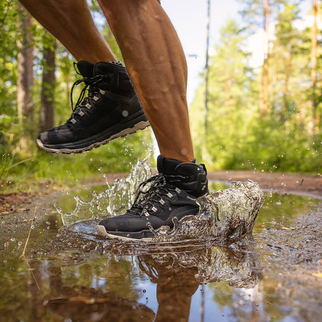 Mustiin vaelluskenkiin pukeutunut henkilö juoksee lätäkön läpi metsäpolulla ja roiskii vettä.