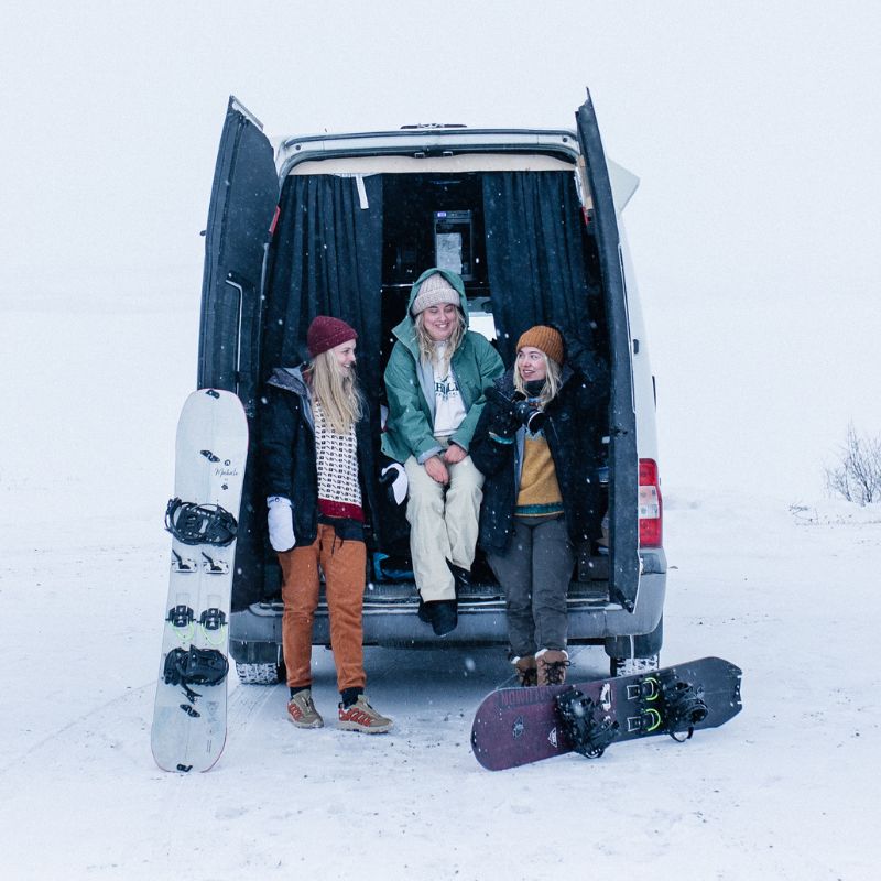 Kolme talvivaatteisiin pukeutunutta ihmistä seisoo ja istuu pakettiauton takapenkillä lumessa, lumilaudat maassa.
