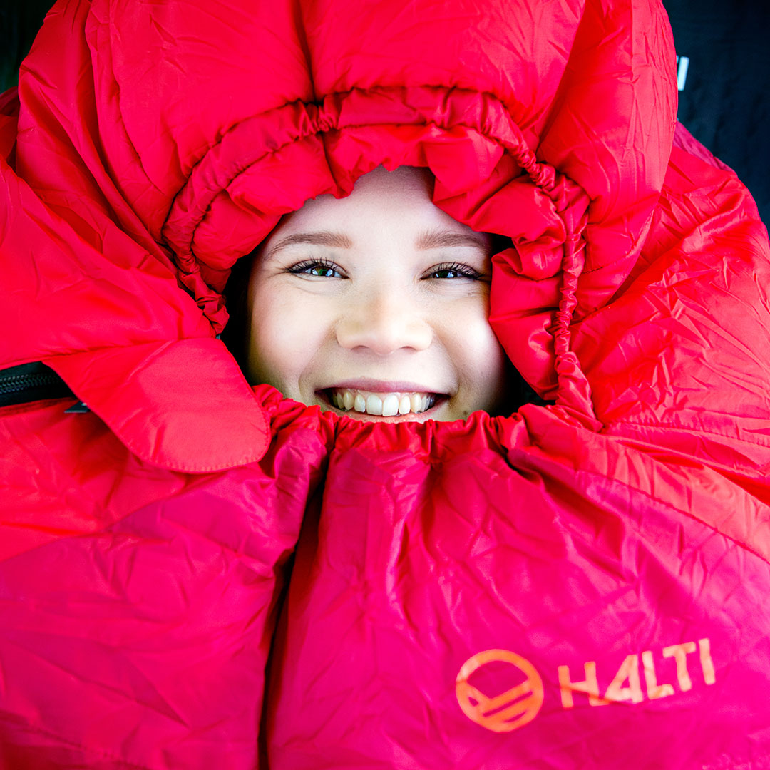 Hymyilevä henkilö, jonka kasvoja kehystää punainen Haltin makuupussi tai eristetakki, katsoo kameraan.