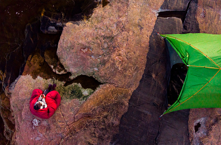 Punaisessa makuupussissa oleva henkilö istuu kalliolla vihreän teltan vieressä ylhäältä katsottuna.