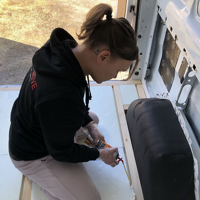 Henkilö polvistuu pakettiauton sisällä ja levittää liimaa pakettiauton lattiaan tiivistyspistoolilla käsineet kädessä.