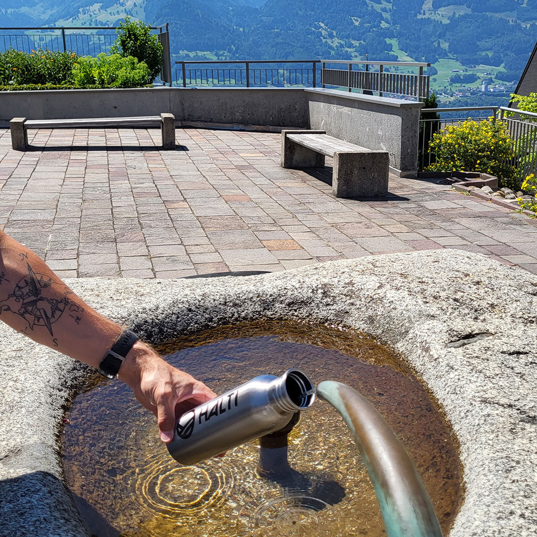 Henkilö täyttää metallisen vesipullon kivilähteestä ulkona, taustalla on vuoria ja penkkejä.