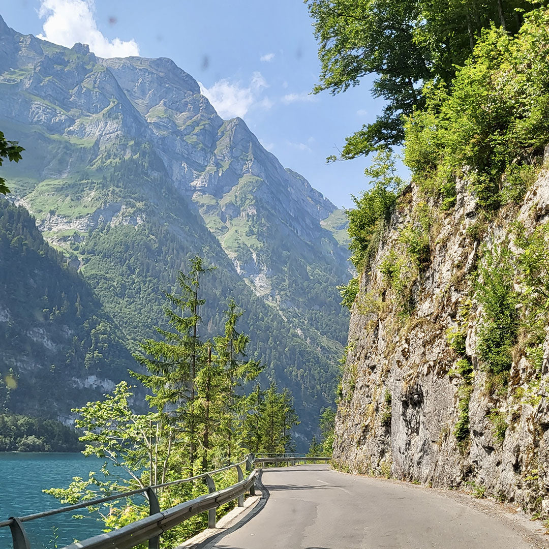 Kapea tie kaartuu kalliojyrkänteen varrella, ja taustalla on vuoria, puita ja sininen järvi kirkkaalla taivaalla.