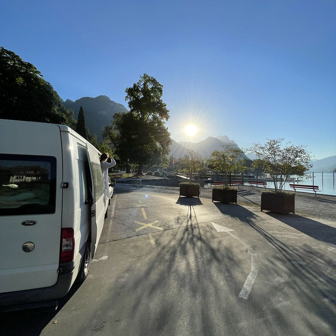 Valkoinen pakettiauto on pysäköity järven läheisyyteen auringonnousun aikaan, taustalla on vuoria ja puita ja maassa on pitkiä varjoja.