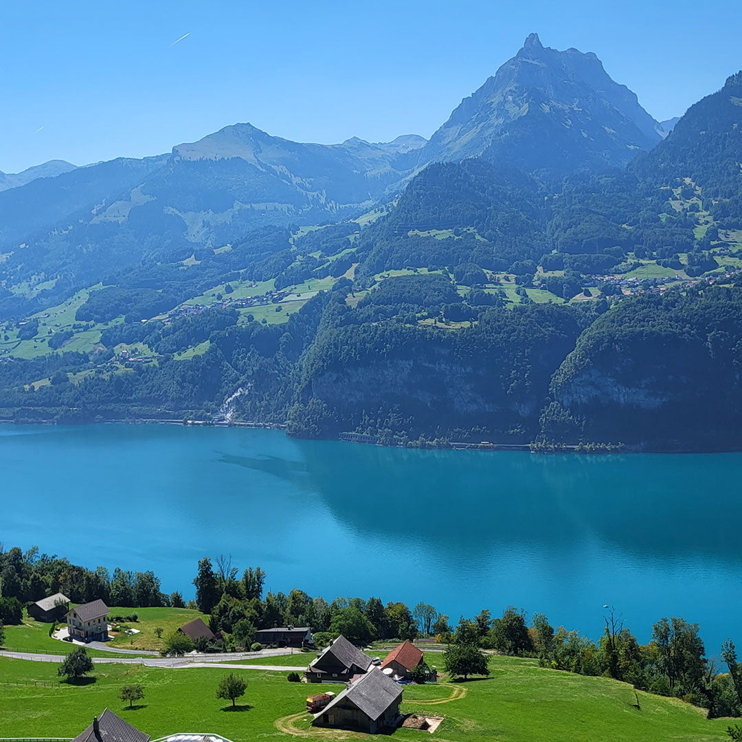 Turkoosi järvi, jonka taustalla on vuoria ja hajallaan olevia taloja nurmikolla sinisen taivaan alla.