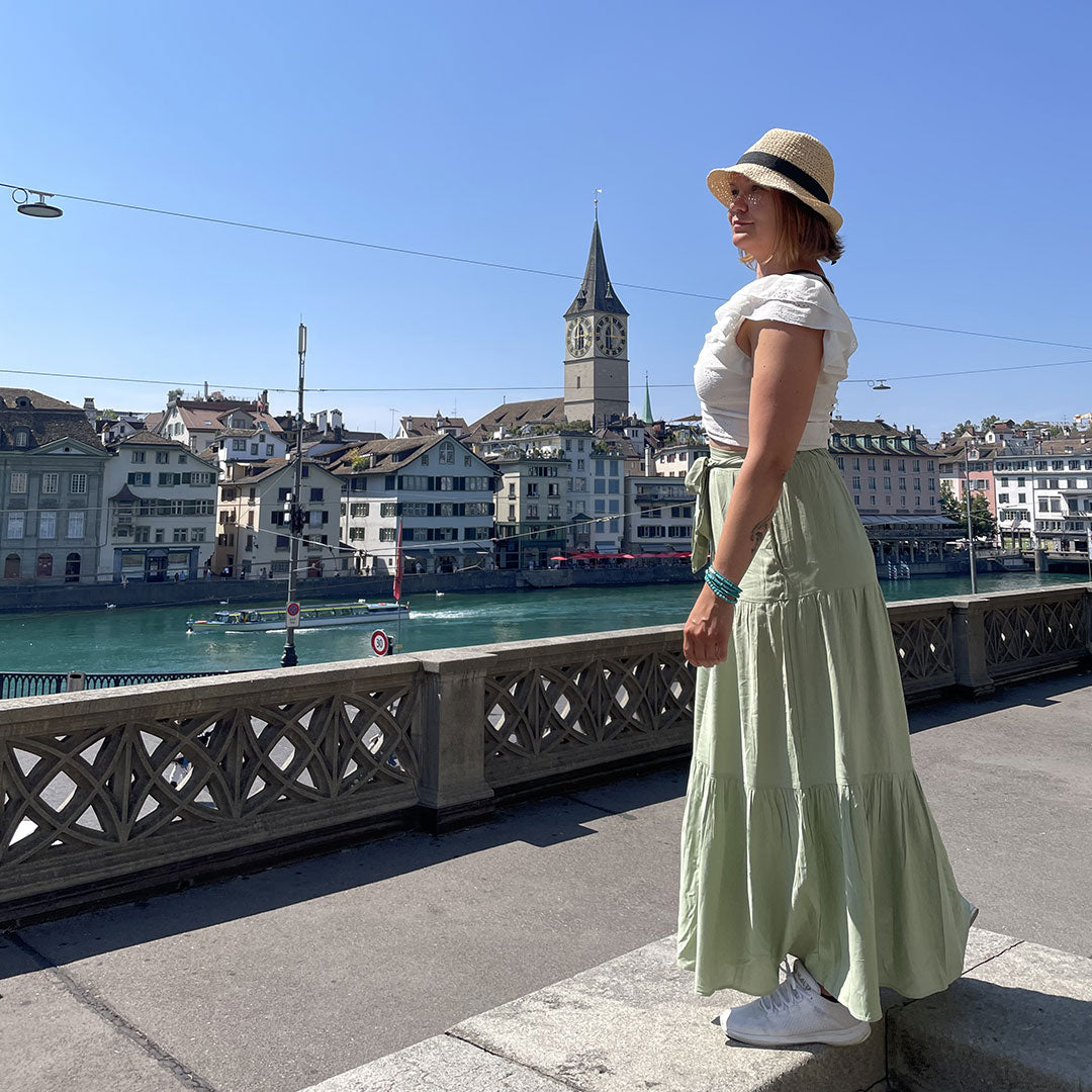 Hattuun ja pitkään hameeseen pukeutunut nainen seisoo joen rannalla kaupungissa, jossa on historiallisia rakennuksia ja kirkon torni.