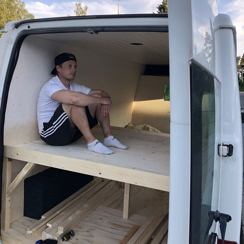 Rennosti pukeutunut mies istuu puisella alustalla osittain muunnetussa pakettiautossa, jonka sivuovi on auki.