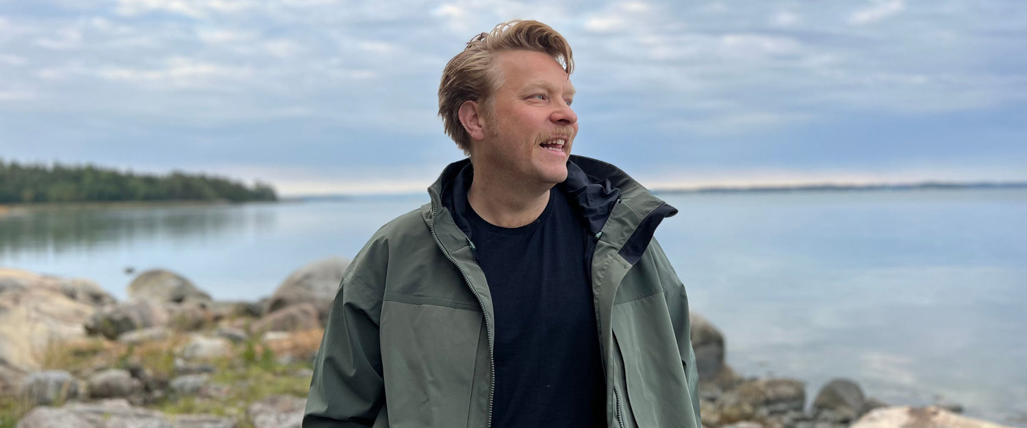 Vihreään takkiin pukeutunut mies seisoo kivikkoisella järven rannalla, jonka taustalla on tyyni vesi ja pilvinen taivas.