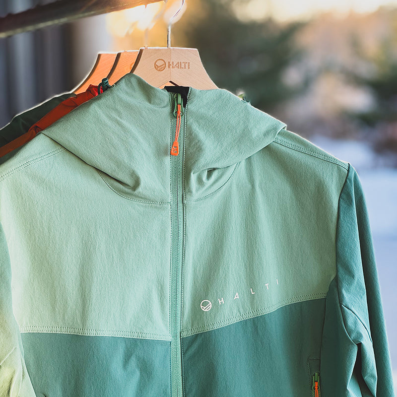 Vaaleanvihreät Halti Pallas Evo Plus X-stretch Takki Naisten -takit roikkuvat ulkona, puut hämärtyvät pehmeästi takana.