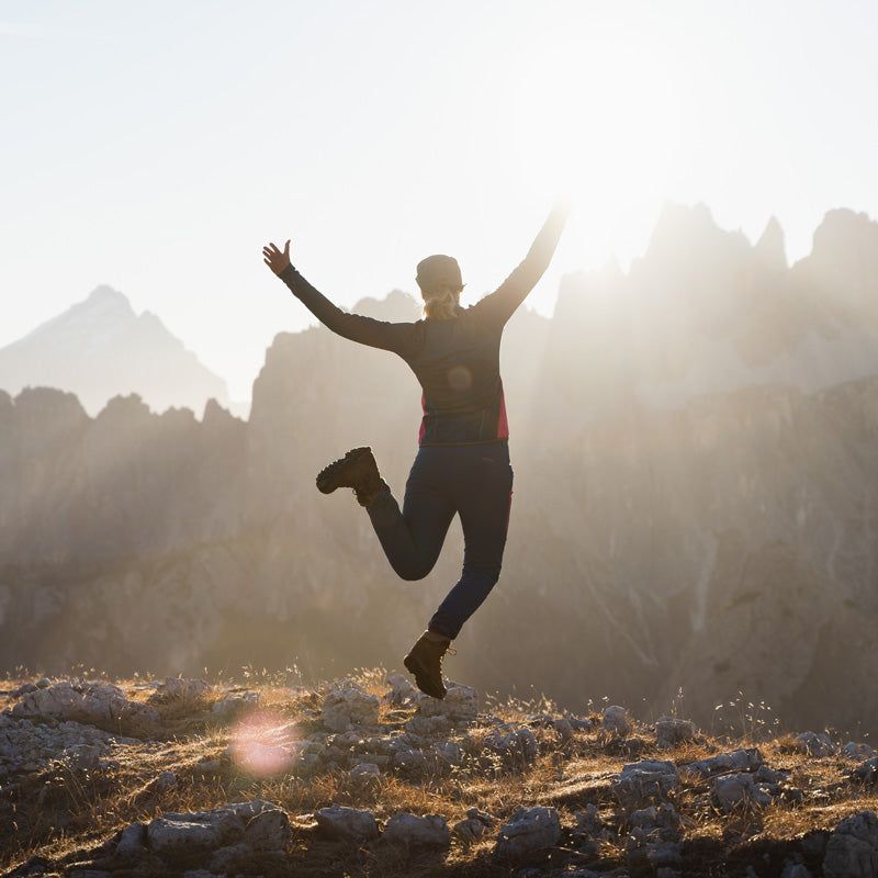 Henkilö seisoo kivikkoisella maalla kädet kohotettuna, siluettina vuoret ja auringonvalo taustalla.
