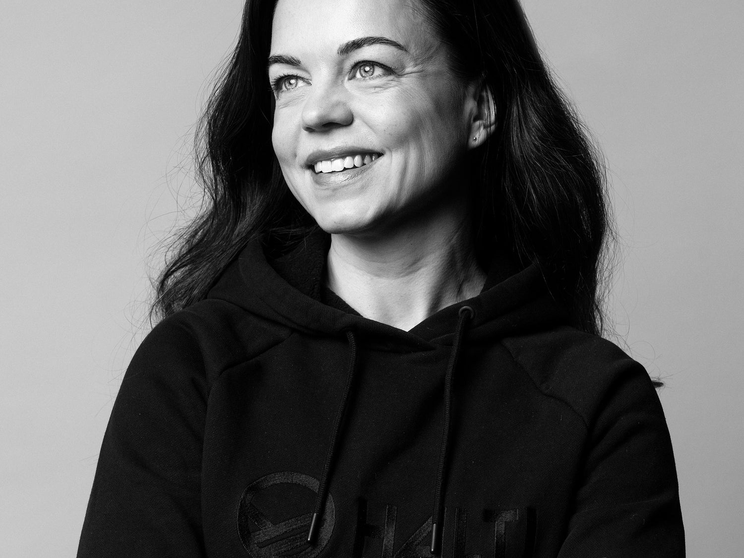 Nainen, jolla on pitkät tummat hiukset, päällään musta huppari, hymyilee ja katsoo hieman sivulle tavallista taustaa vasten.