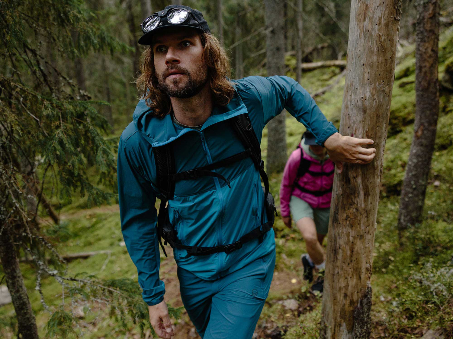 Siniseen takkiin pukeutunut mies vaeltaa metsässä ylämäkeen, ja toinen vaaleanpunaiseen takkiin pukeutunut henkilö kävelee hänen takanaan.