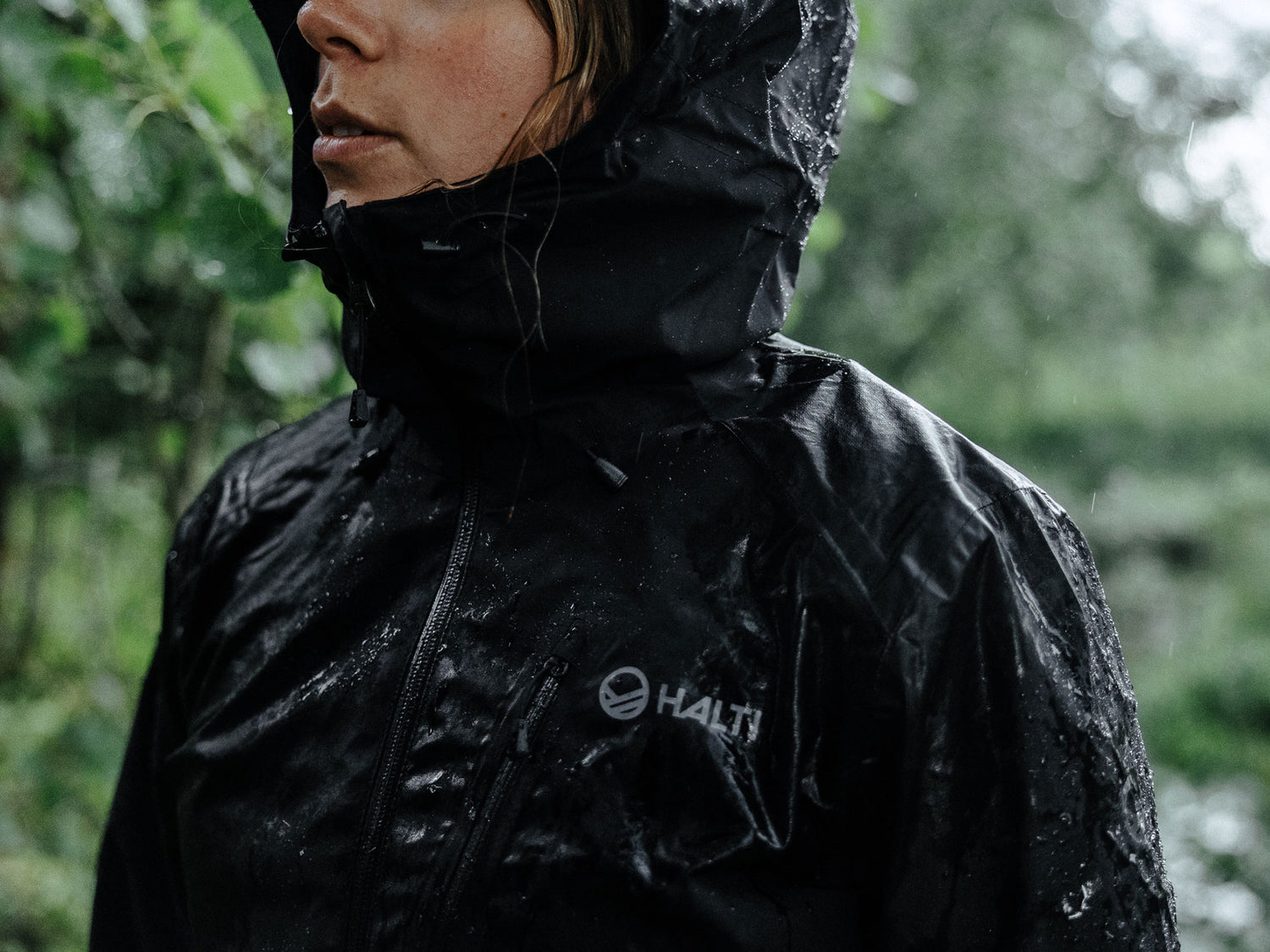 Henkilö, jolla on musta Halti-sadetakki ja huppu ylhäällä, seisoo ulkona sateessa ja takin päällä on vesipisaroita.