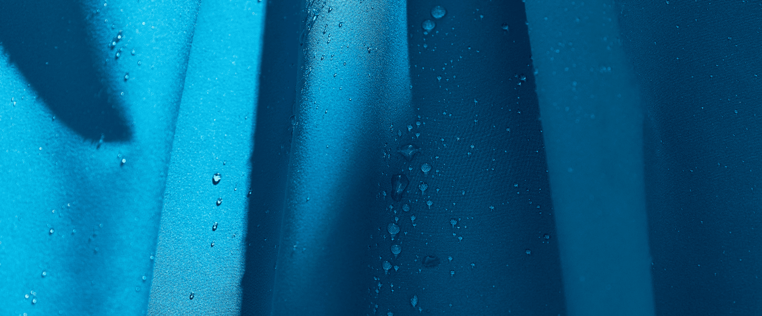 Lähikuva sinisestä kankaasta, jonka pinnalle on ripoteltu vesipisaroita, jotka luovat valon ja varjon alueita.