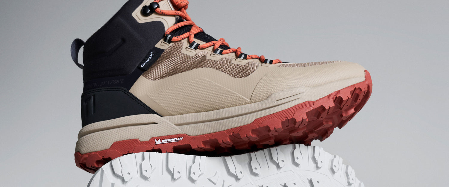 Beigen ja mustan värinen vaelluskenkä, jossa on punaiset nauhat ja punainen karhea pohja, joka lepää valkoisella kuvioidulla pinnalla.